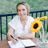 Kasia smiling and sitting outside on her deck at table with sunflowers while writing in her journal