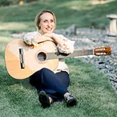 Kasia sitting on the grass outside holding her guitar with her arms crossed while resting on top of the guitar